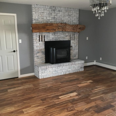 Modern living room with a gray brick fireplace and dark firebox, wood mantel, gray accent wall, crystal chandelier above, and warm wood plank flooring.